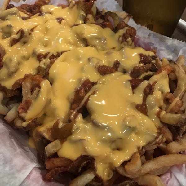 Close-up of golden french fries topped with chili and melted yellow cheese, served in a paper-lined basket.