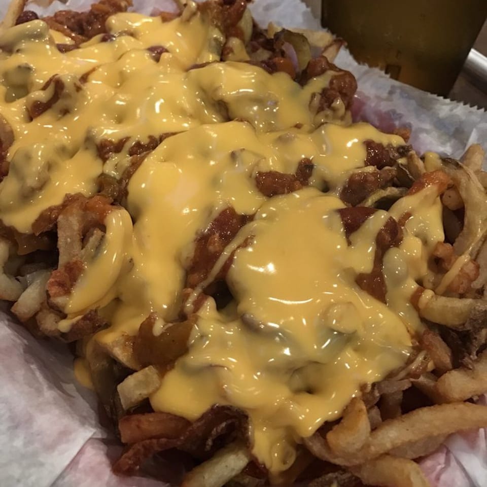 Close-up of golden french fries topped with chili and melted yellow cheese, served in a paper-lined basket.